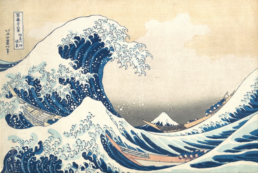 Woodblock print: a huge blue wave, its crest breaking into fingers of white foam, curls over three long, narrow boats whose rowers crouch low, while the small snow-capped cone of Mount Fuji sits in the distance under a pale sky. A title cartouche and the signature are at the top left.