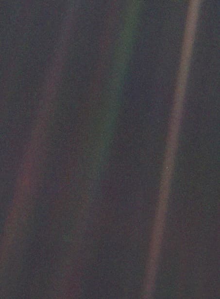 Photograph: a dark field crossed by faint bands of scattered sunlight in pink, green and brown. In the brightest band, on the right, sits one tiny pale speck: the Earth.